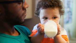 Father helping baby drink bottle in kitchen